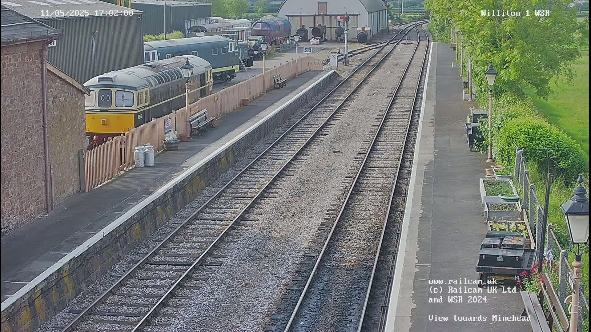 A diesel locomotive sits at a railway station platform on a partly sunny day, with multiple railway tracks extending into the distance.
