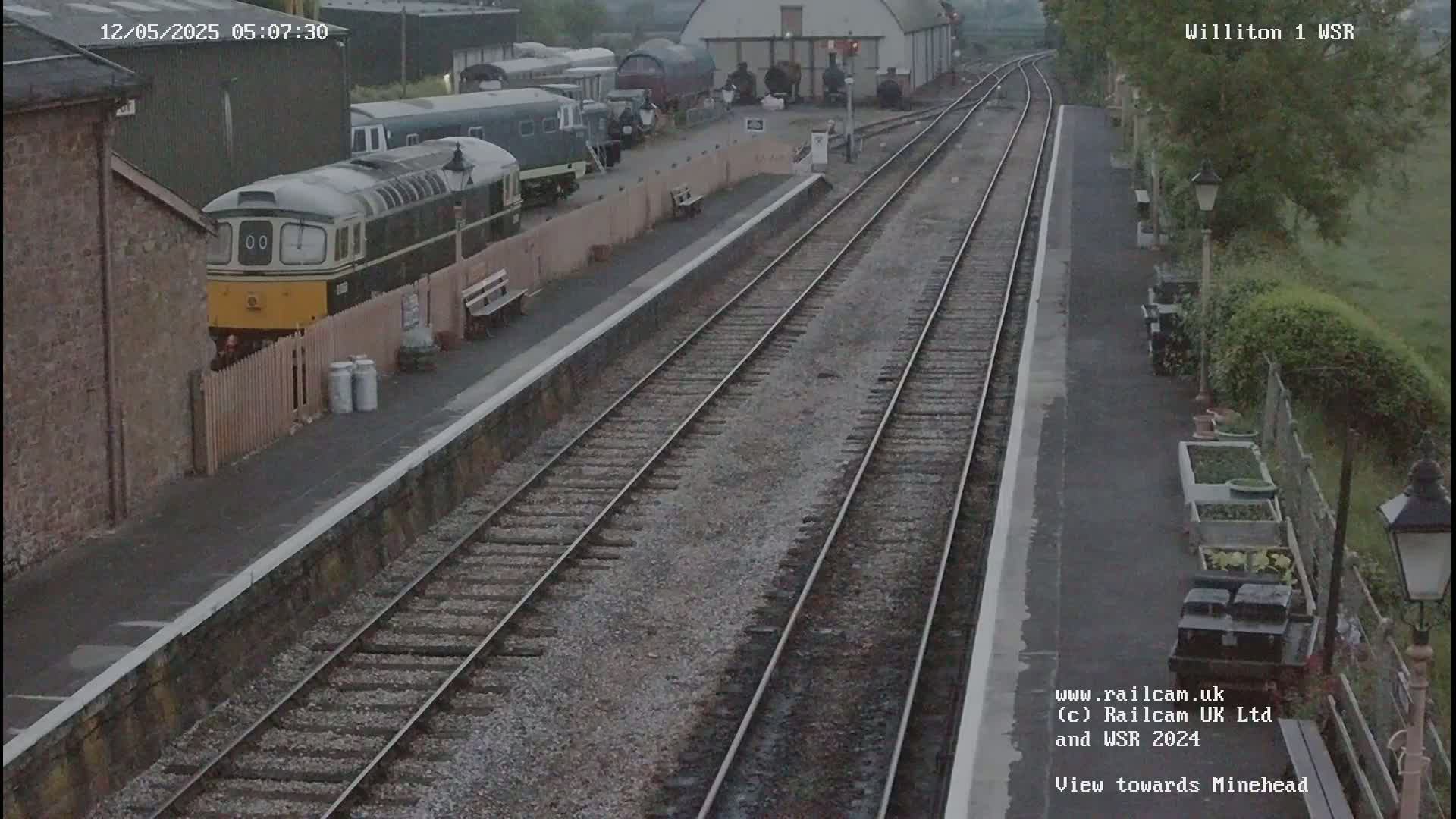 A diesel locomotive sits at a train station platform next to several railway cars under an overcast sky.