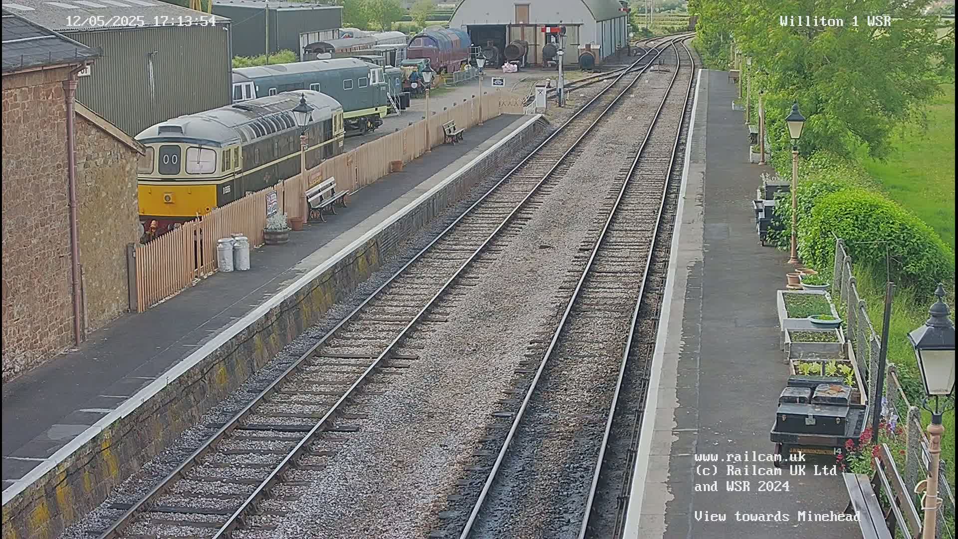 A diesel locomotive sits at a train station platform next to multiple train cars and railway tracks on a partly sunny day.