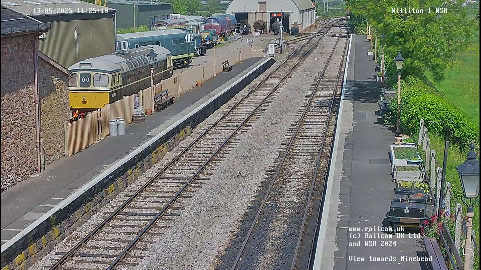 A sunny day at a train station shows a yellow and green diesel locomotive parked next to a platform with multiple train tracks extending into the distance.