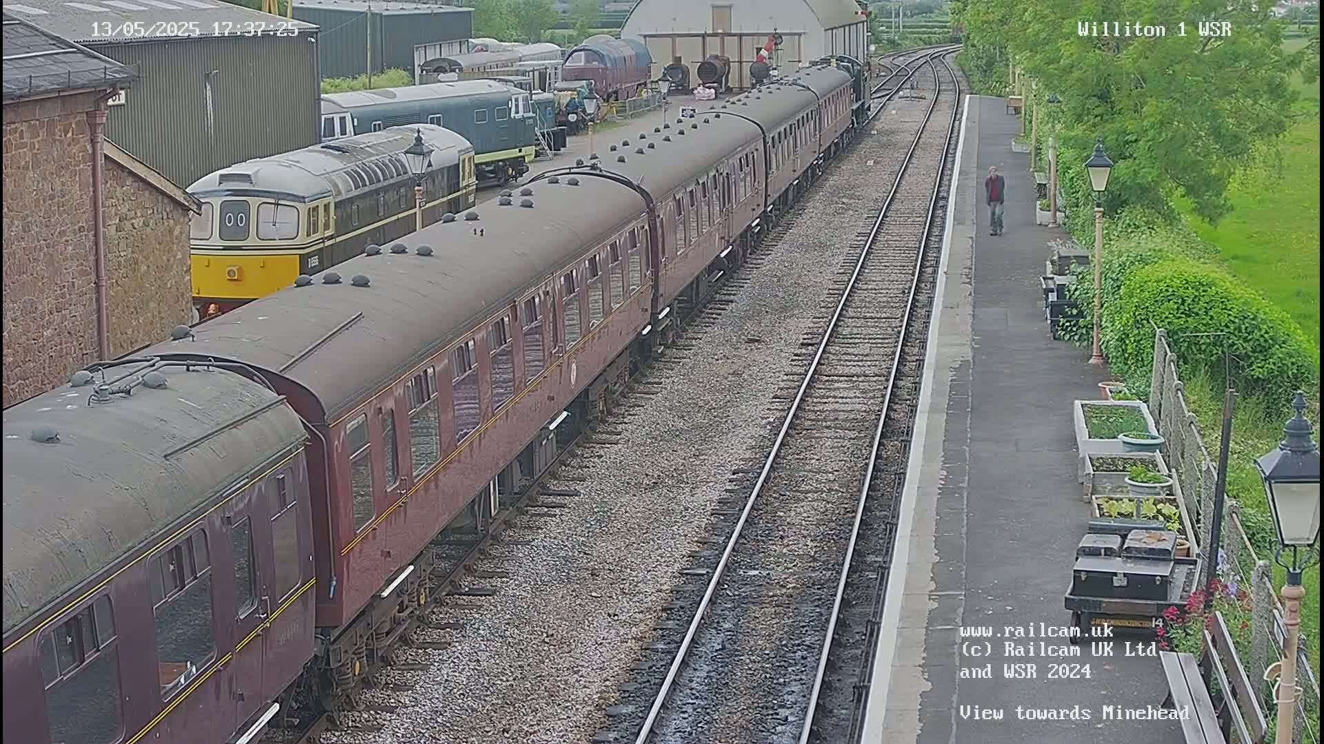 A long maroon passenger train sits at a station on a partly cloudy day, with a diesel locomotive at its head and another train visible in the background.