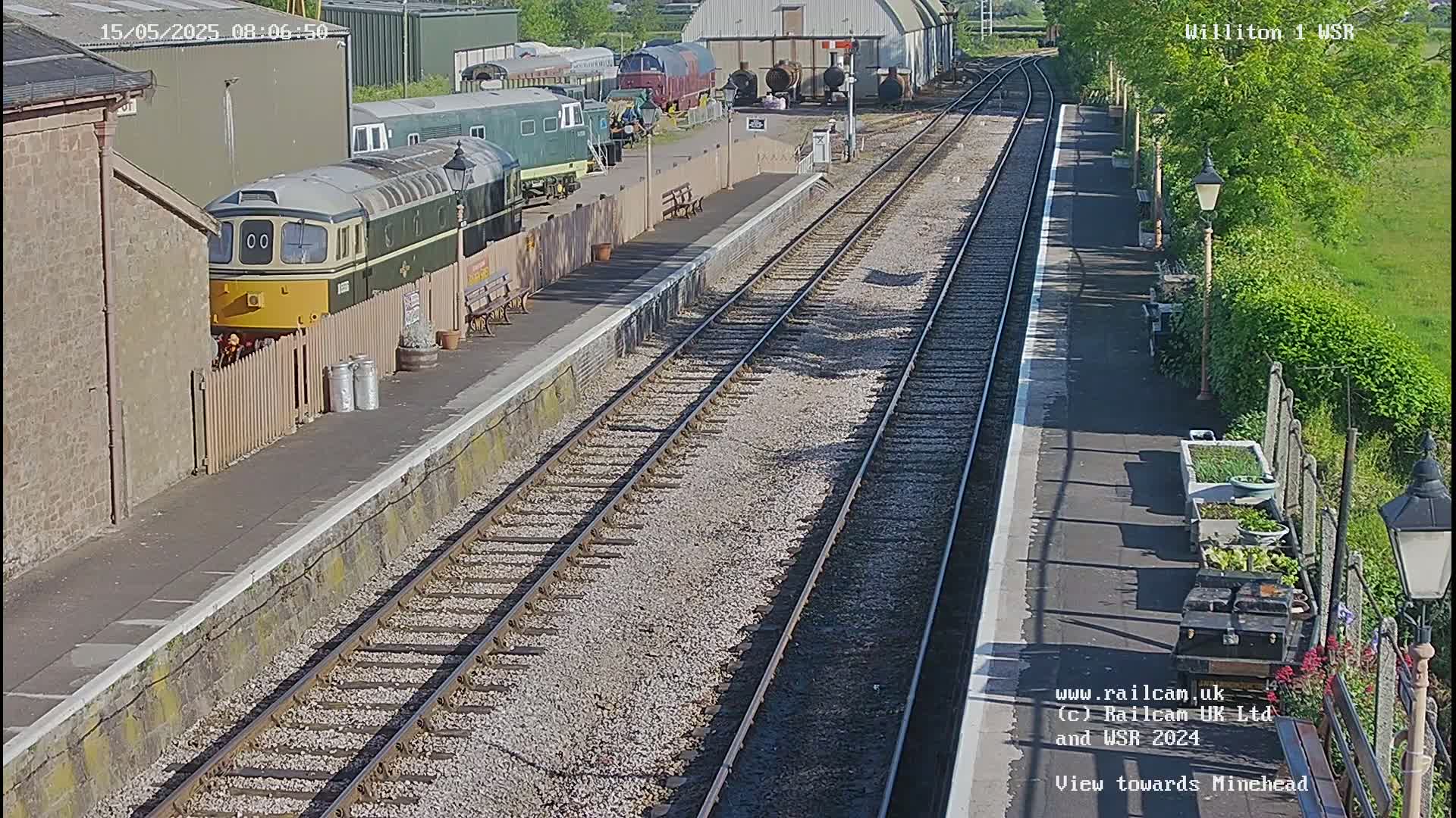 A sunny view of a train station with a diesel locomotive and several railway carriages parked on a platform, alongside multiple train tracks extending into the distance.