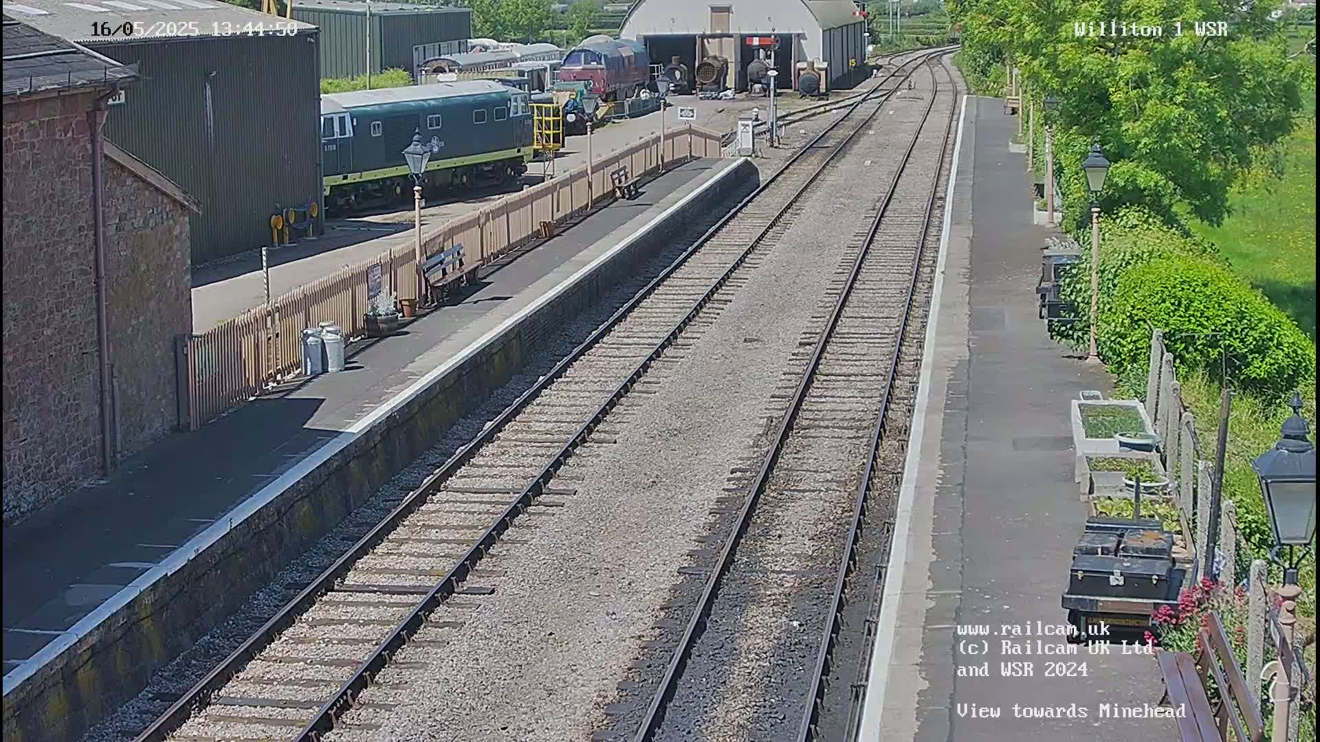 A sunny day at a train station shows multiple train tracks leading to a platform with several parked trains and buildings nearby.