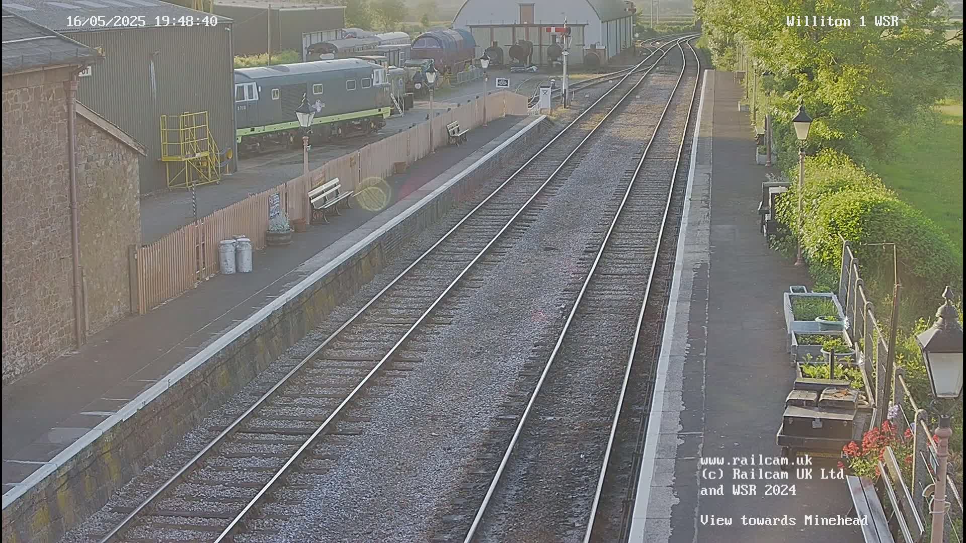 A train station platform with two train tracks, a diesel train visible in the background, and a partially sunny day.
