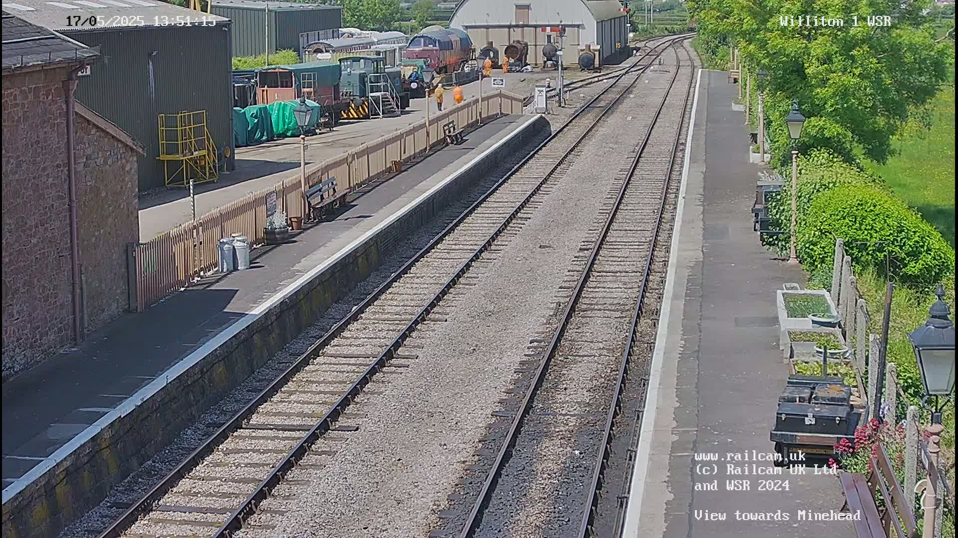 A sunny day at a train station shows multiple train cars parked beside a platform with railway tracks extending into the distance.