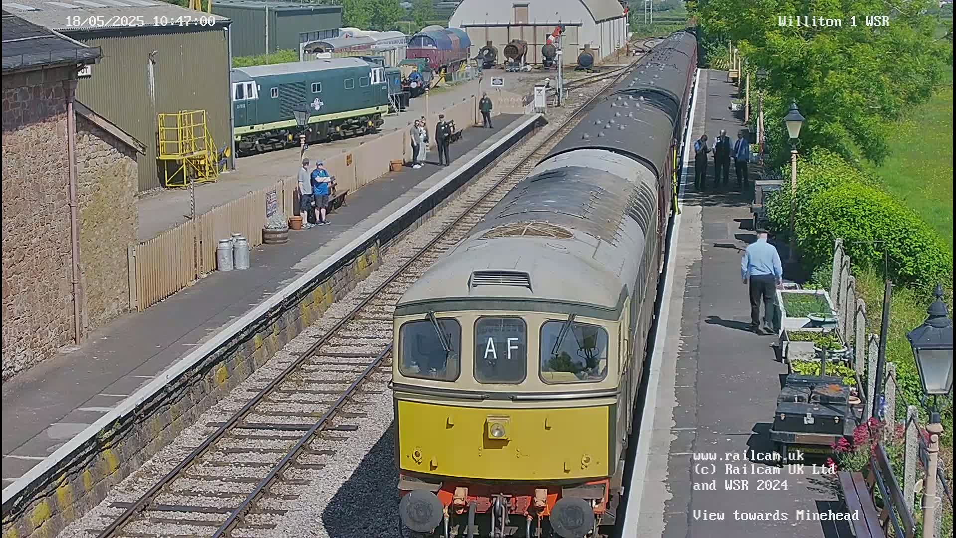 A diesel train is at a station platform on a sunny day, with other trains and people visible nearby.