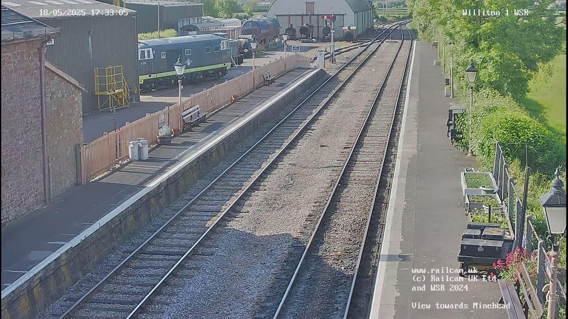 A train station platform on a sunny day with multiple train tracks, a parked locomotive, and adjacent buildings.
