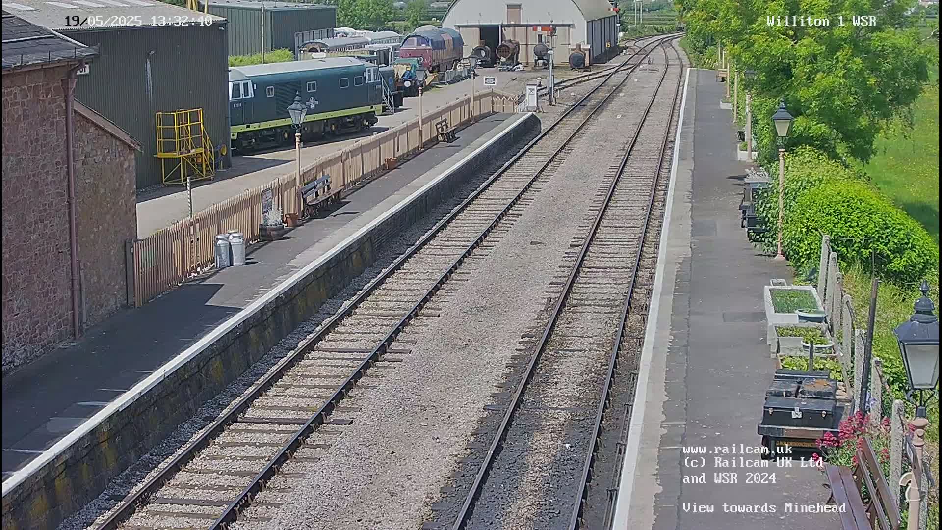 A sunny day at a train station shows multiple train tracks extending into the distance, with several trains and train cars parked nearby.