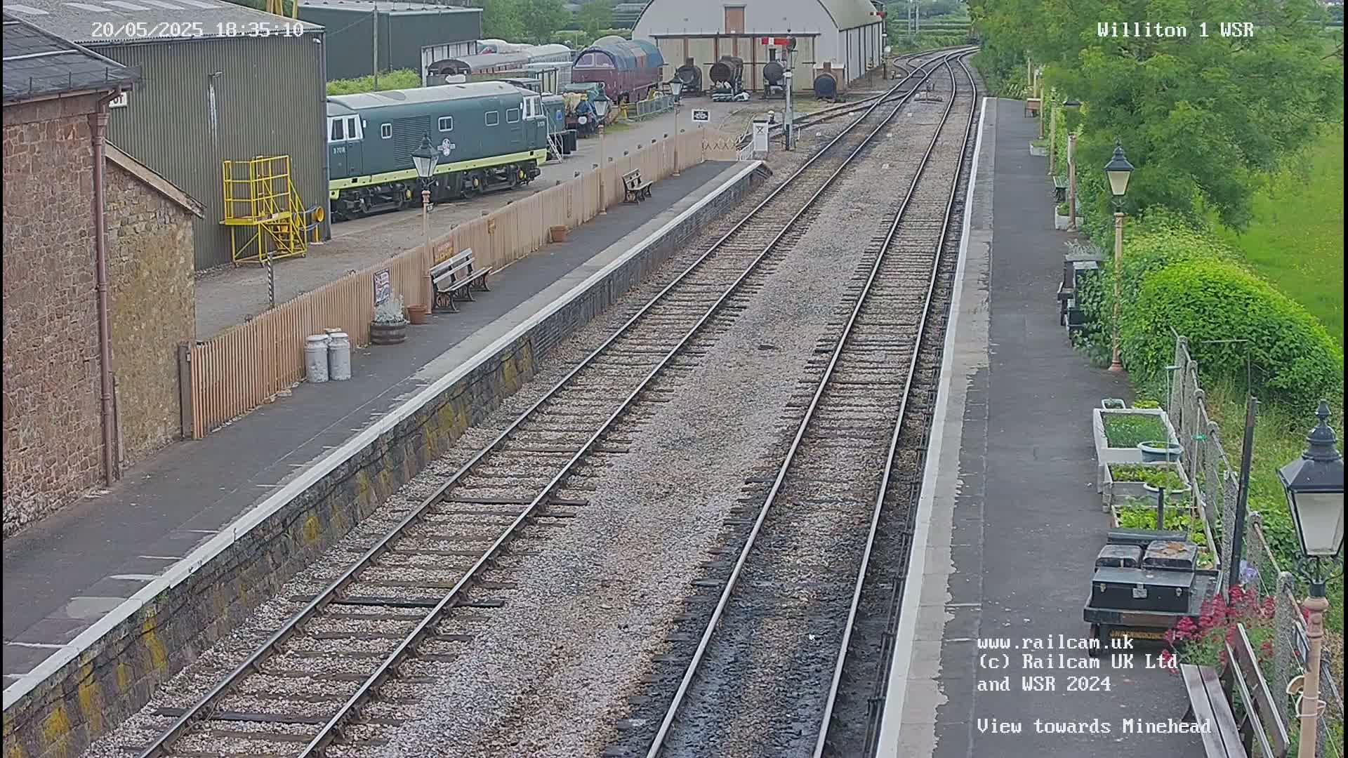 A train station platform with multiple train tracks, a dark-green diesel locomotive, and some railway buildings is visible on a partly sunny day.