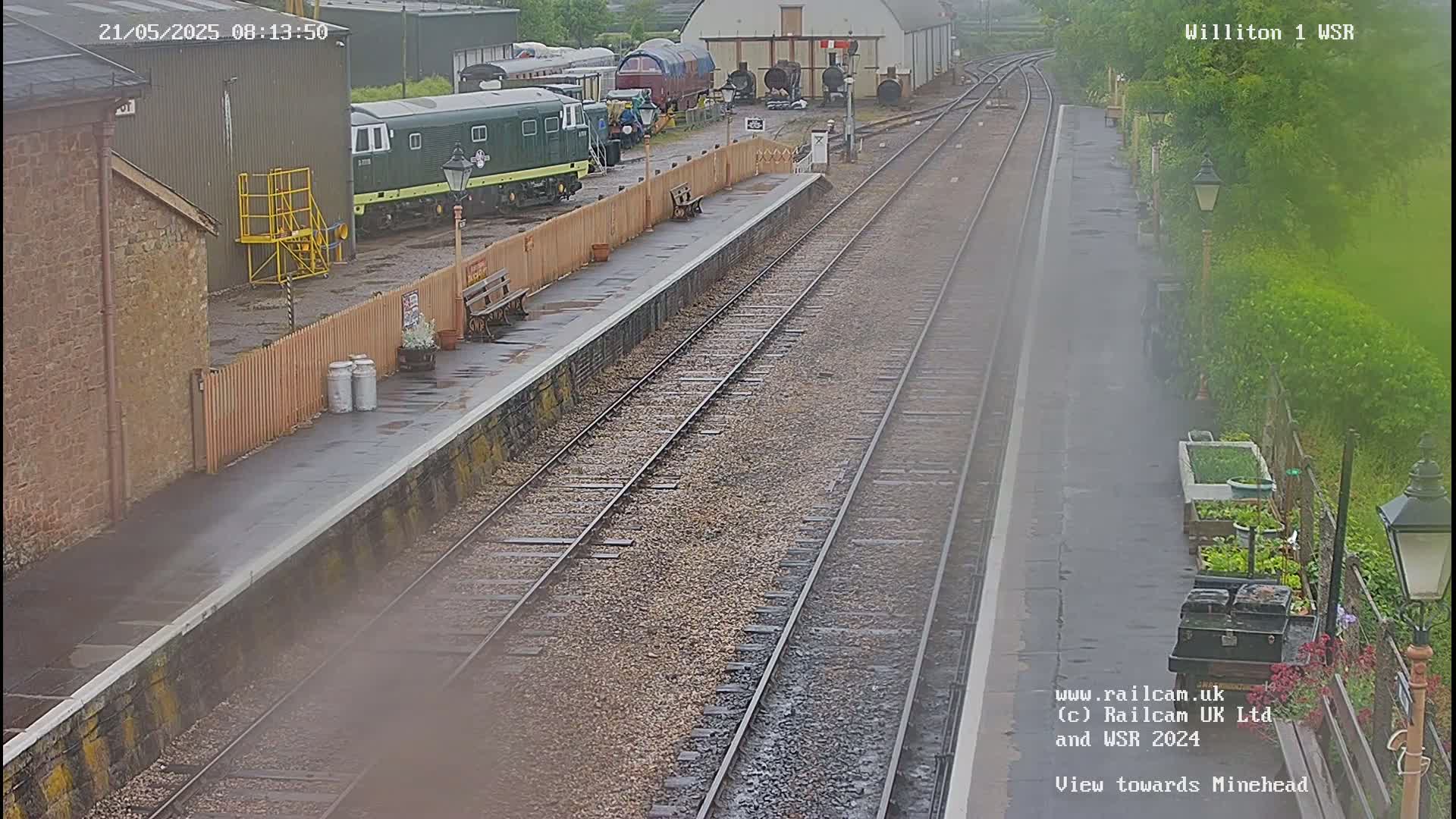 A wet train station platform with several railway tracks, train cars, and a building in the background.