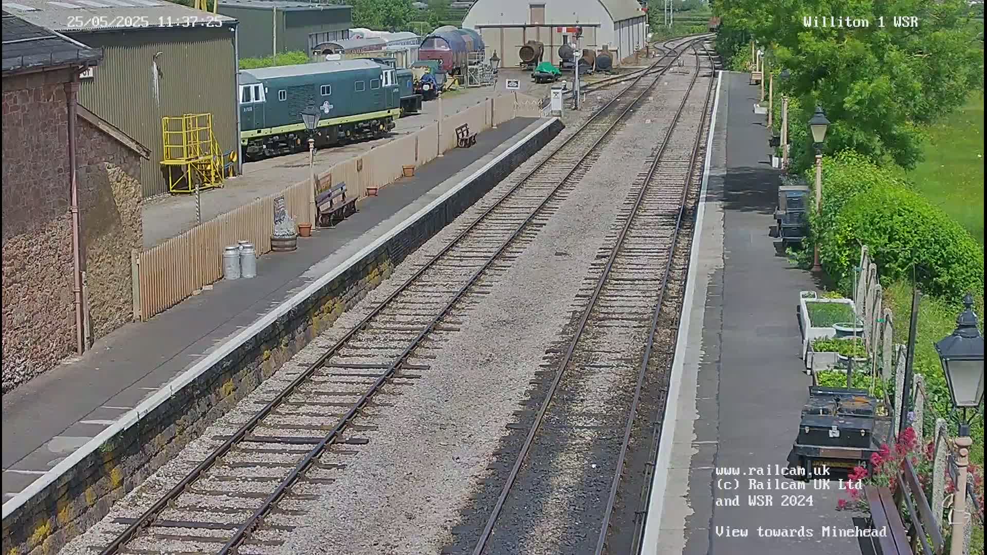 A railway station platform with two parallel train tracks, a green diesel locomotive parked nearby, and a sunny day.