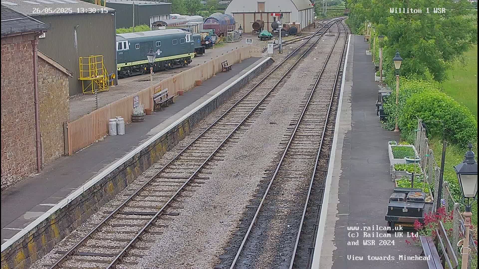 A train station platform with several railway tracks, a green diesel locomotive parked nearby, and a small garden alongside the platform on a partly cloudy day.