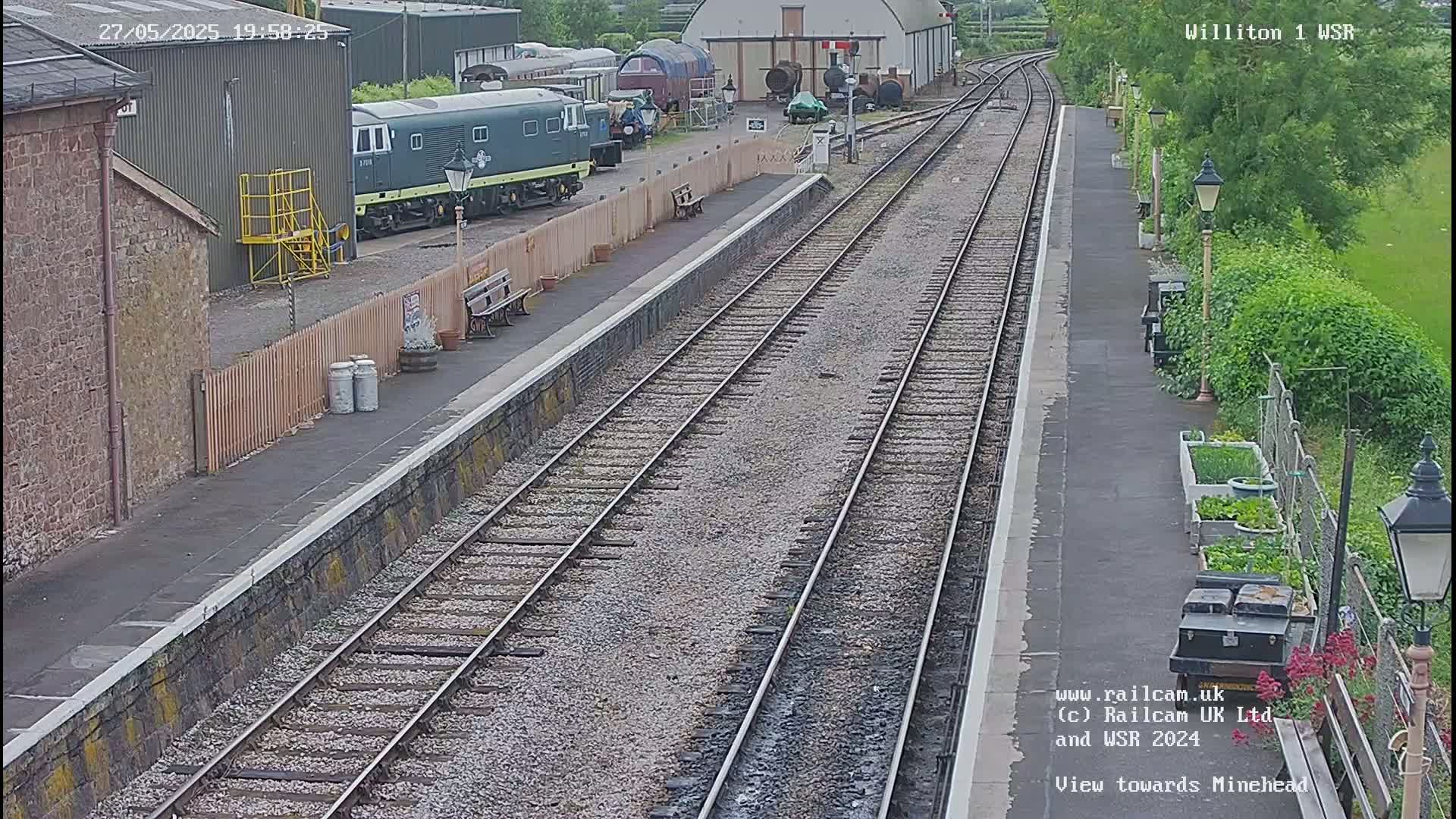 A railway station platform with two train tracks, a diesel locomotive parked nearby, and a small garden beside the tracks on an overcast day.