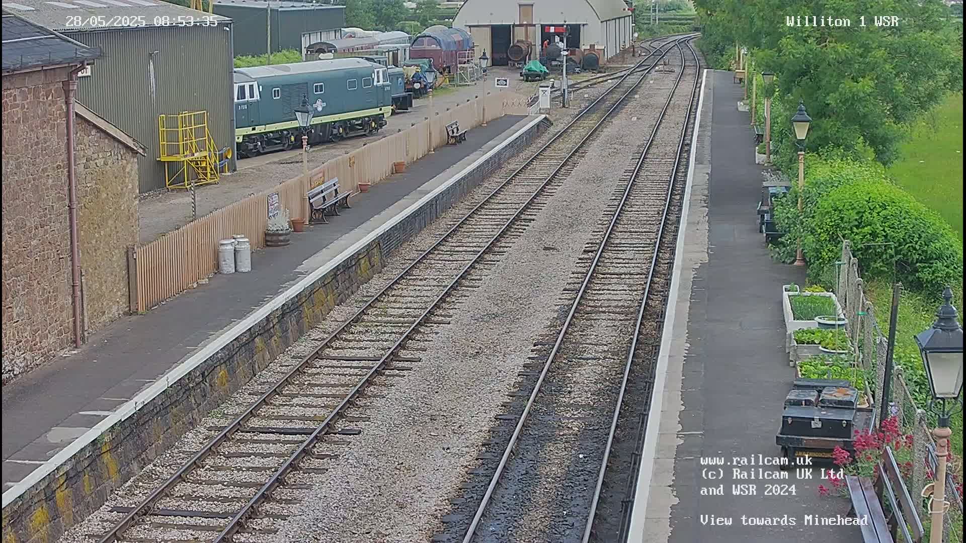 A train station platform with multiple train tracks extending into the distance on a partly cloudy day.