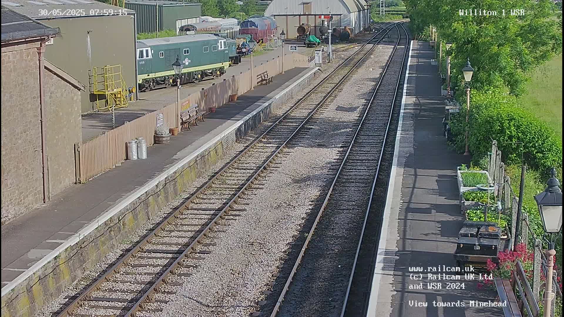A sunny view of a train station platform with multiple train tracks, a green diesel locomotive parked nearby, and a small garden area.