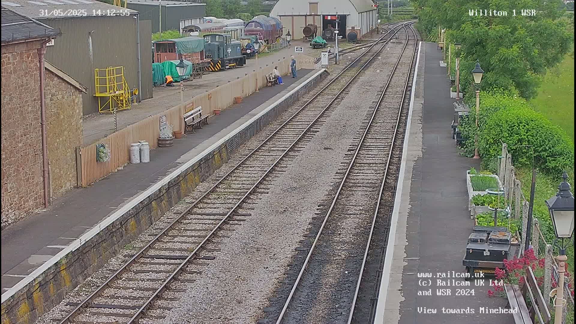 A railway platform with multiple train tracks, several buildings, and a small garden, on a partly sunny day.