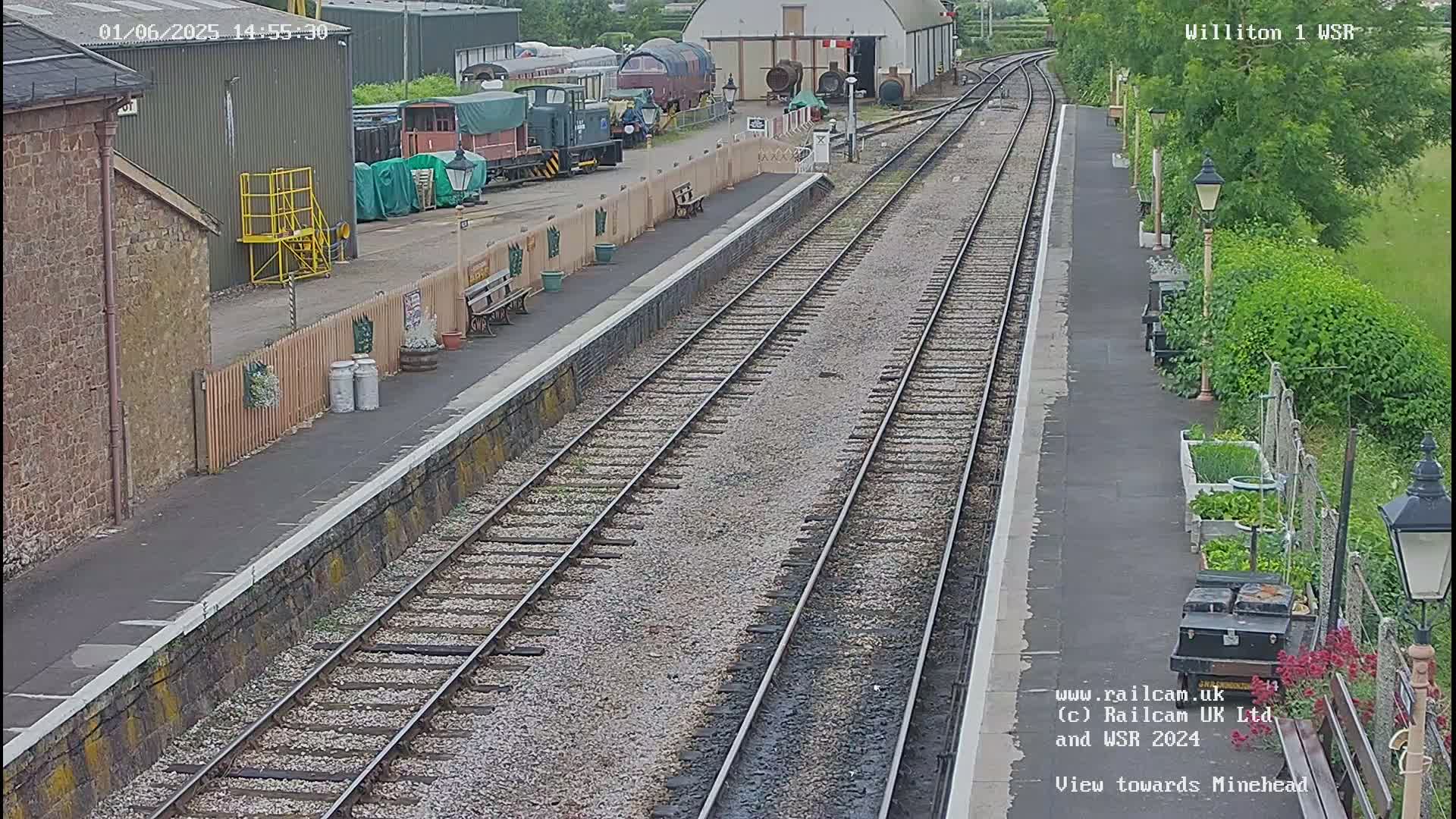 A train station platform with multiple train tracks, railway buildings, and a small garden is shown on an overcast day.