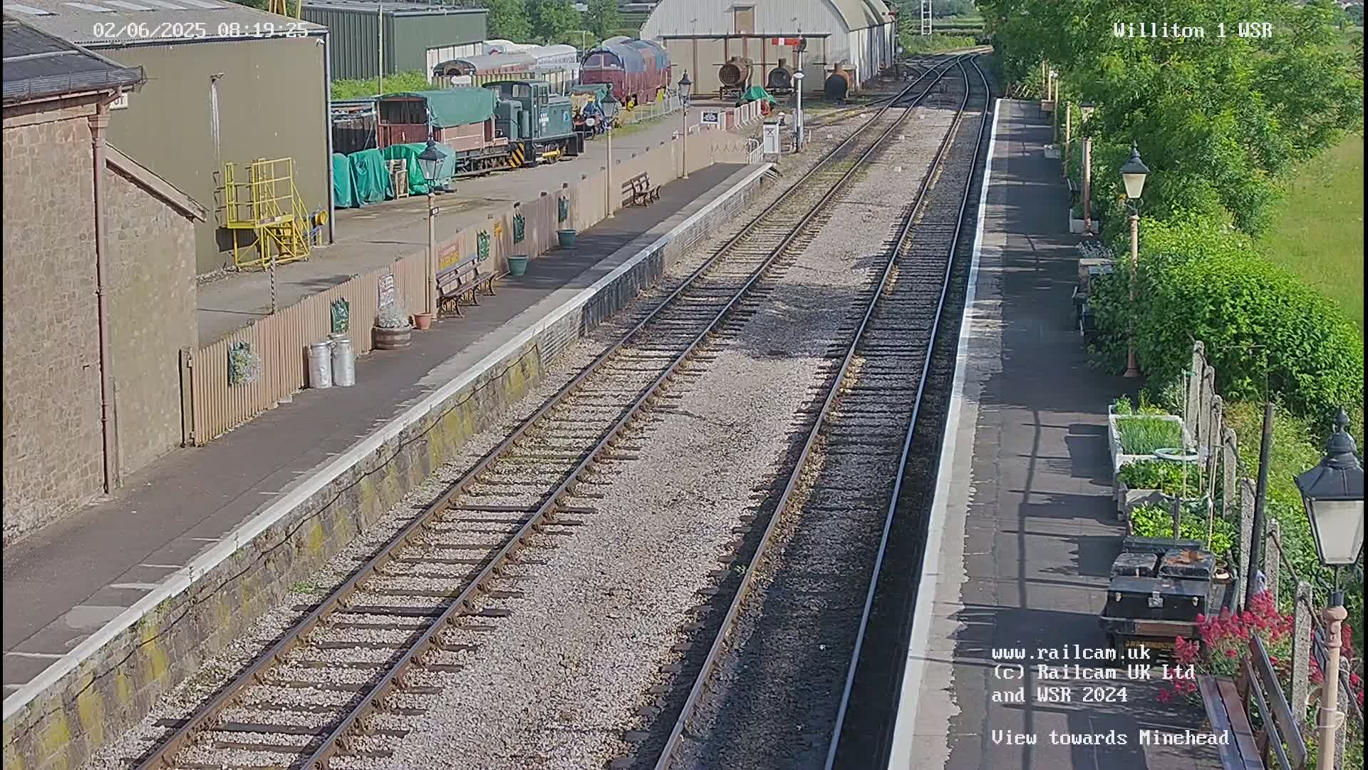 A sunny day at a train station shows two parallel train tracks extending into the distance, with several railway cars parked to the left, and a platform with benches and a small garden to the right.