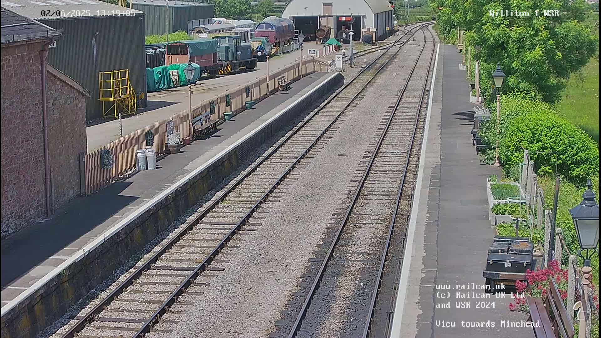 A sunny view of a railway station platform with multiple train tracks, railway cars, and a building in the background.