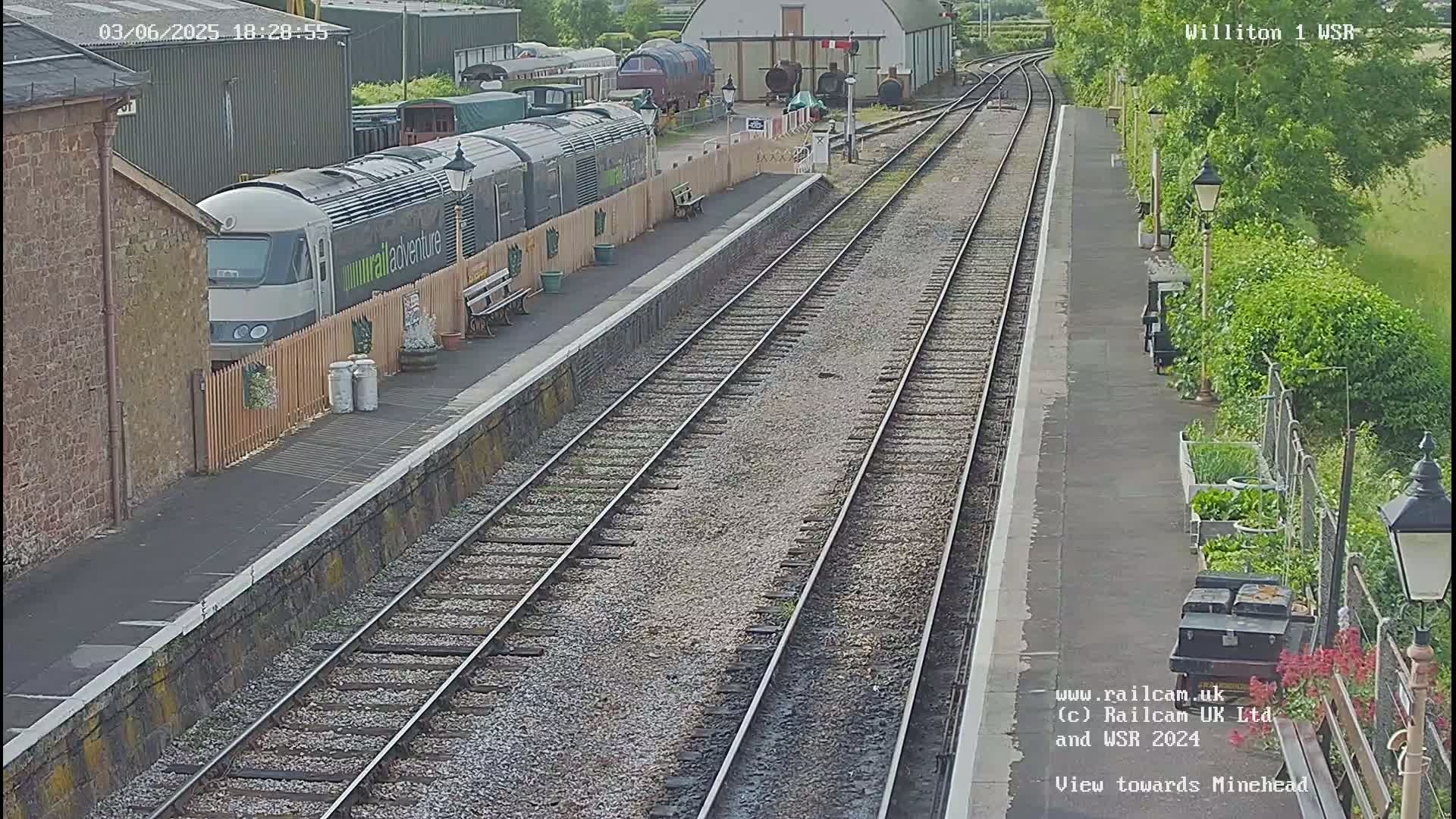 A long train is parked at a station platform next to a fenced area, with multiple train tracks extending into the distance on a sunny day.