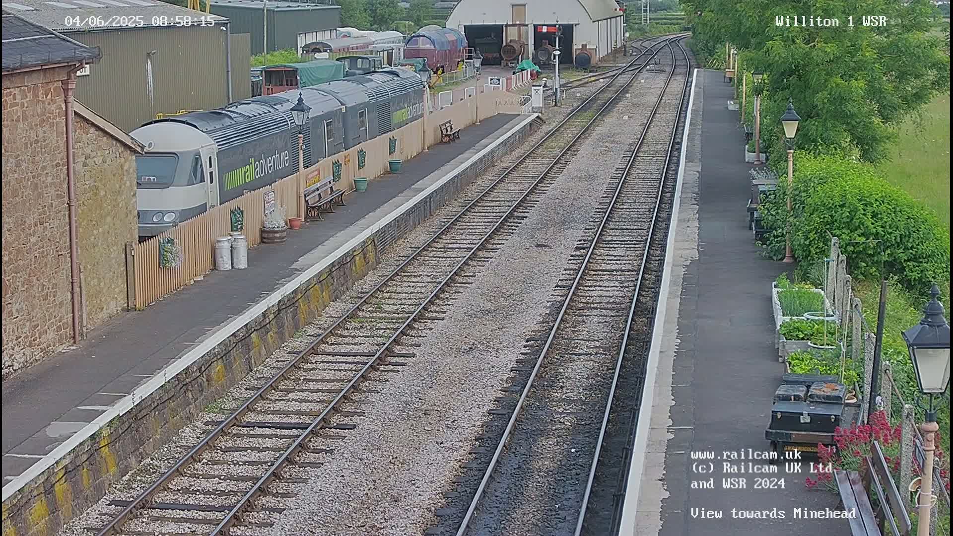 A long train sits at a station platform next to a building, with railway tracks extending into the distance on a partly cloudy day.