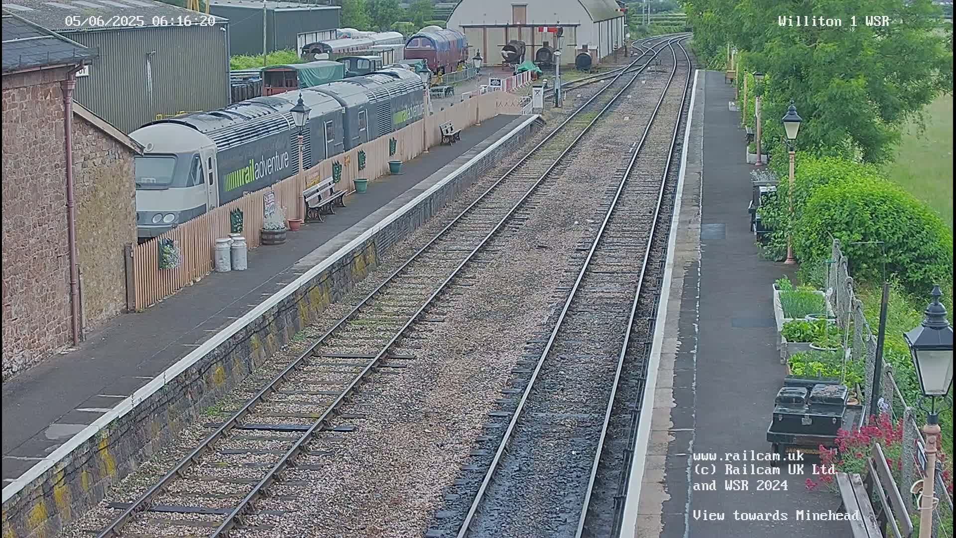 A train is parked at a railway station platform on a partly cloudy day, with multiple train tracks extending into the distance.
