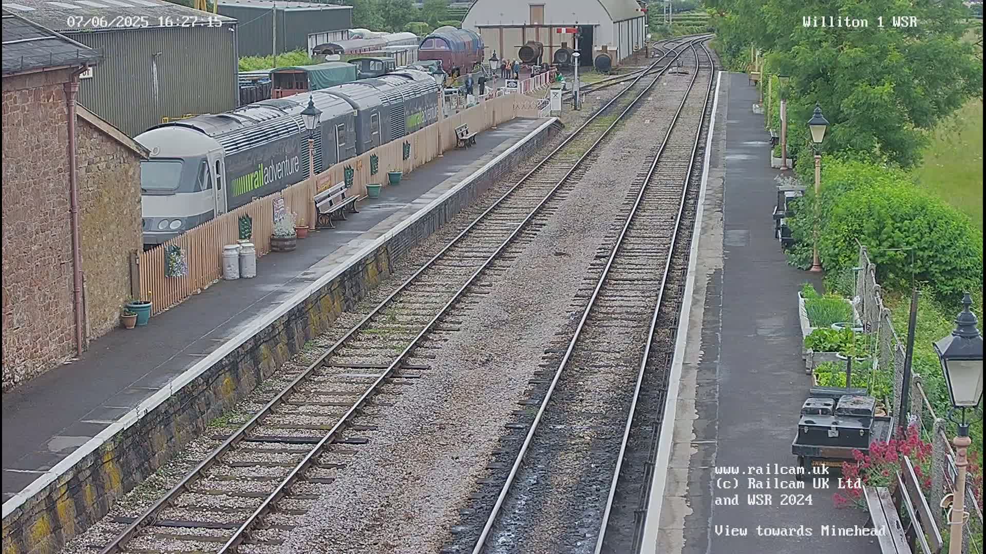 A long train is parked at a station platform next to several railway tracks, under a partly sunny sky.