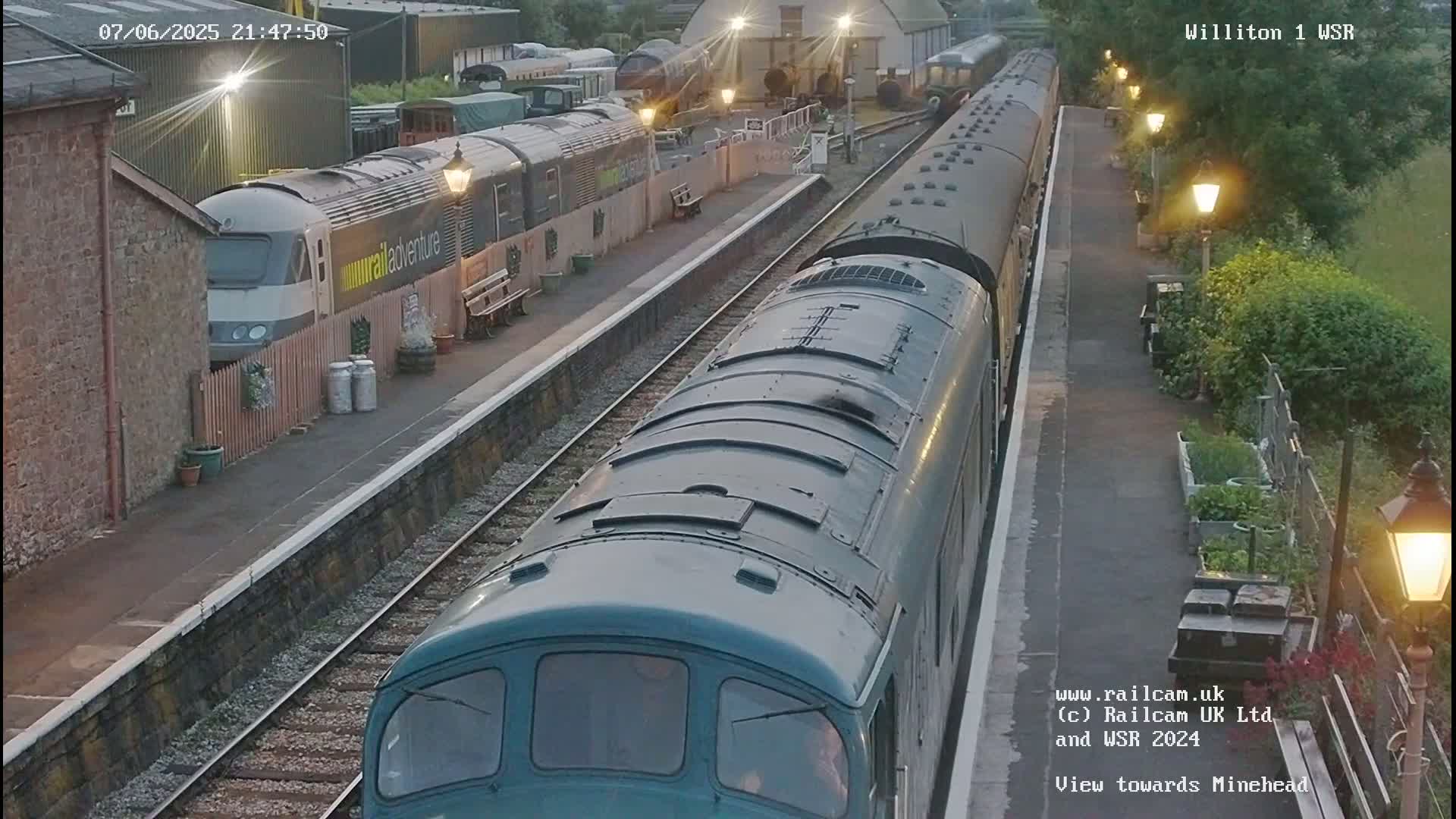 A teal diesel locomotive and a long passenger train are at a station under a twilight sky, with additional train cars parked nearby.