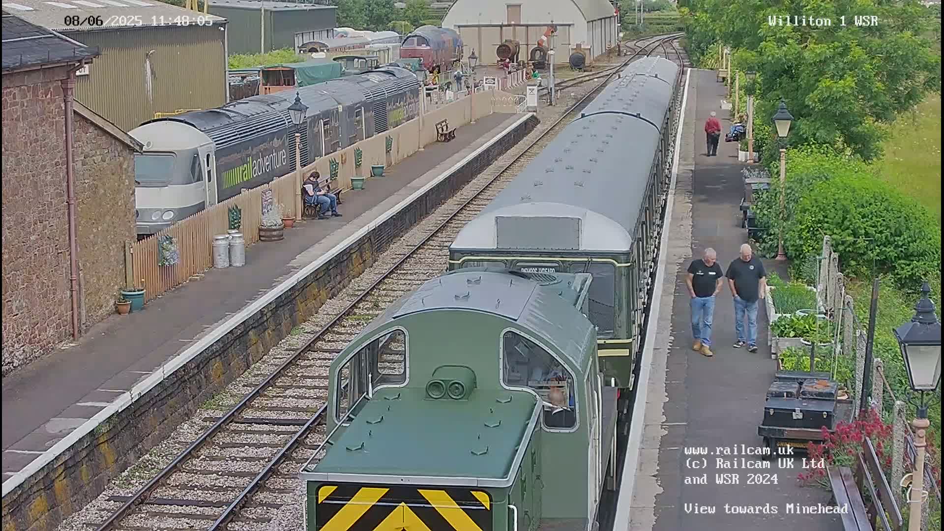A train station with two trains, one green and one gray, on the tracks on a partly sunny day.