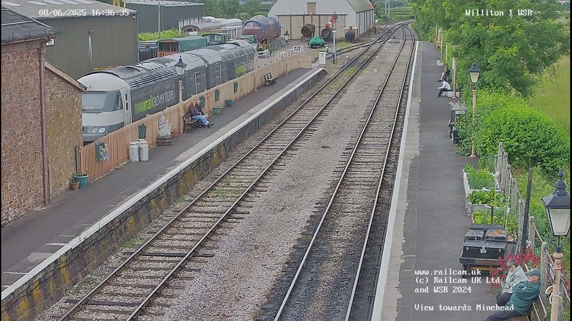 A train sits at a station platform on a partly sunny day, with several people sitting on benches nearby.