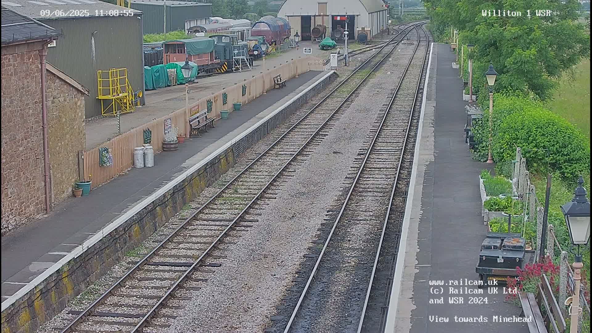 A view down a railway track at a train station on an overcast day, with railway cars and buildings visible in the background.