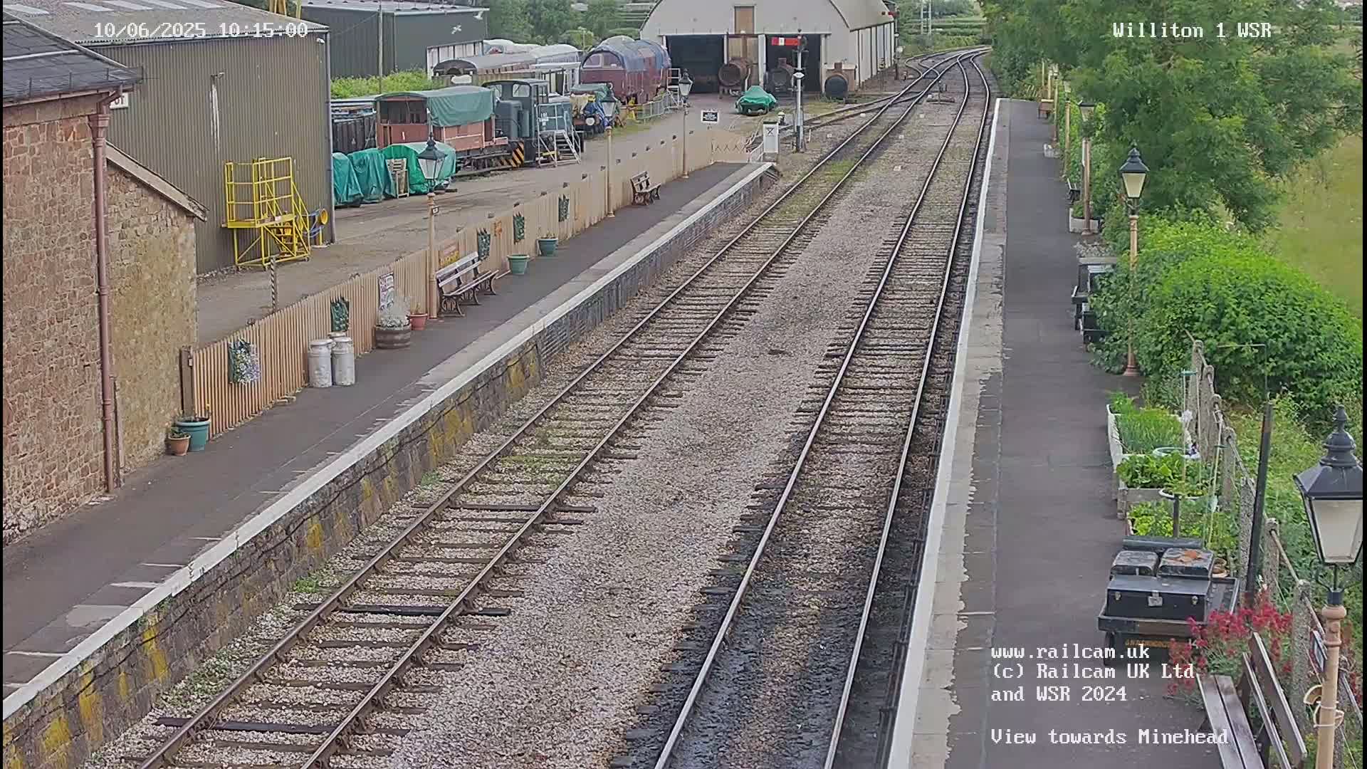 A train station platform with multiple train tracks extends into the distance on a partly cloudy day, with several railway cars visible nearby.
