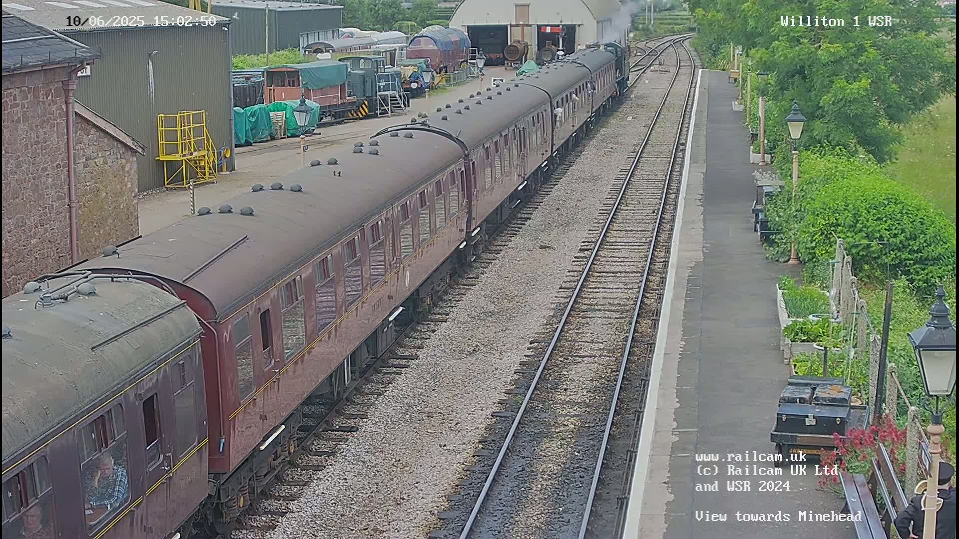 A long maroon passenger train sits on a railway track next to a platform with a small garden beside it on a partly sunny day.