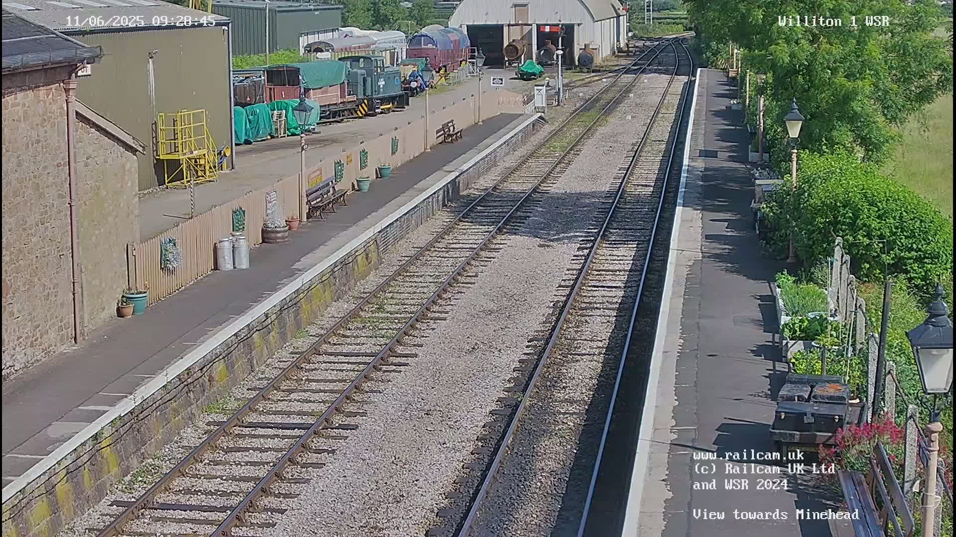 A sunny view of a train station platform with several train cars and a building visible in the background.