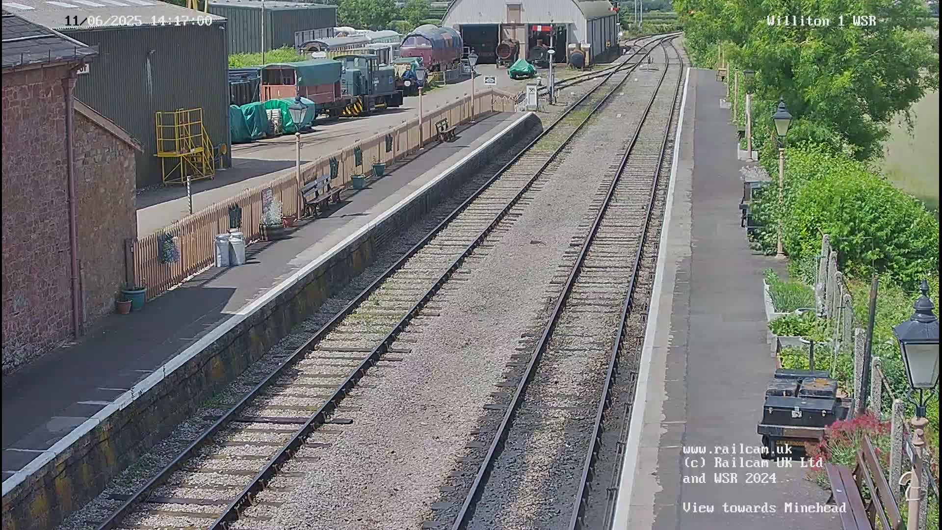 A sunny view of a train station platform with railway tracks extending into the distance, several railway cars visible in the background, and some landscaping along the side of the platform.
