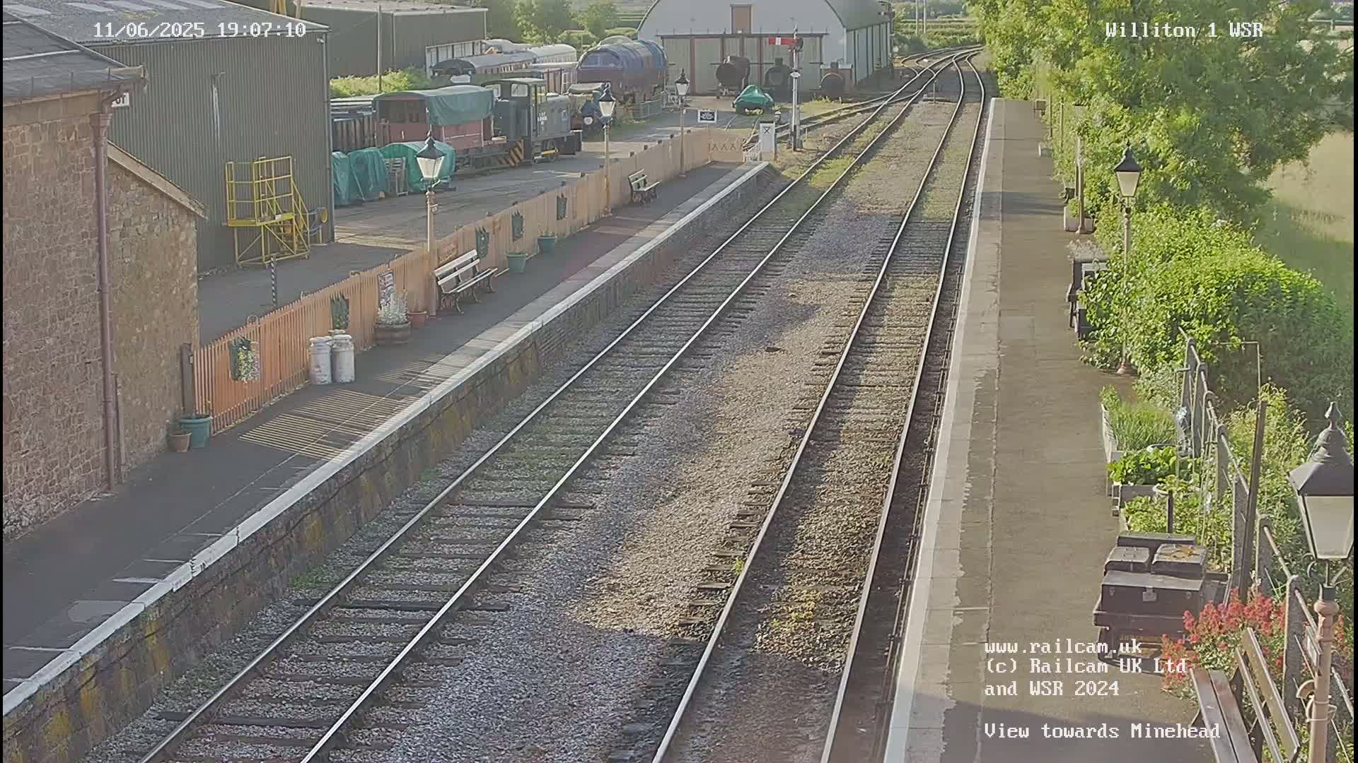 A train station platform with multiple train tracks, several railway cars visible in the background under a sunny sky.