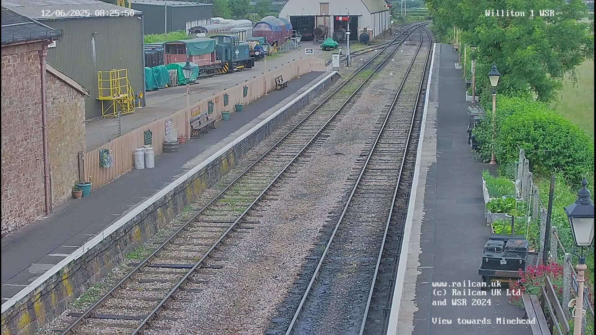 A train station platform with multiple train tracks, several railway carriages, and a small garden beside it on a partly cloudy day.