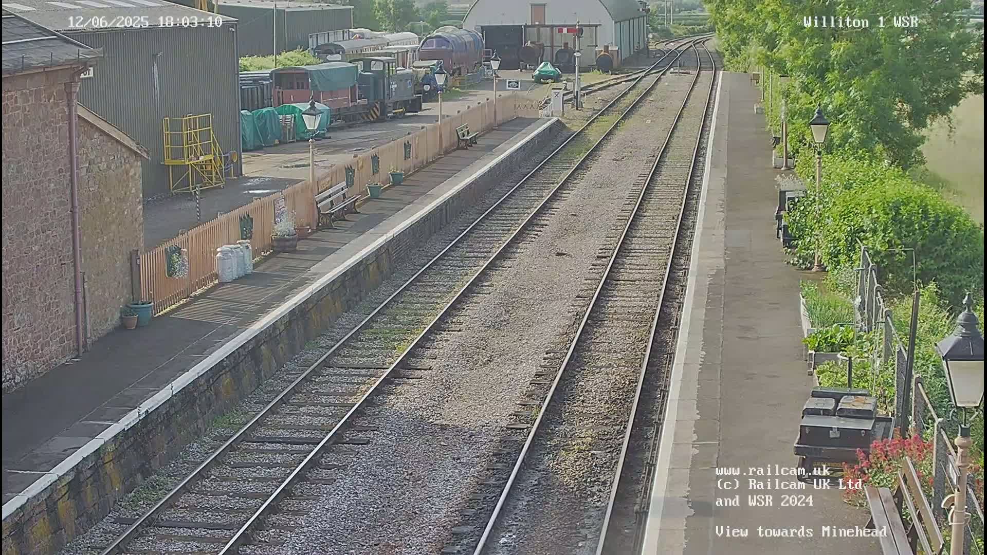 A train station with several train cars visible, under sunny conditions, shows two parallel train tracks extending into the distance.