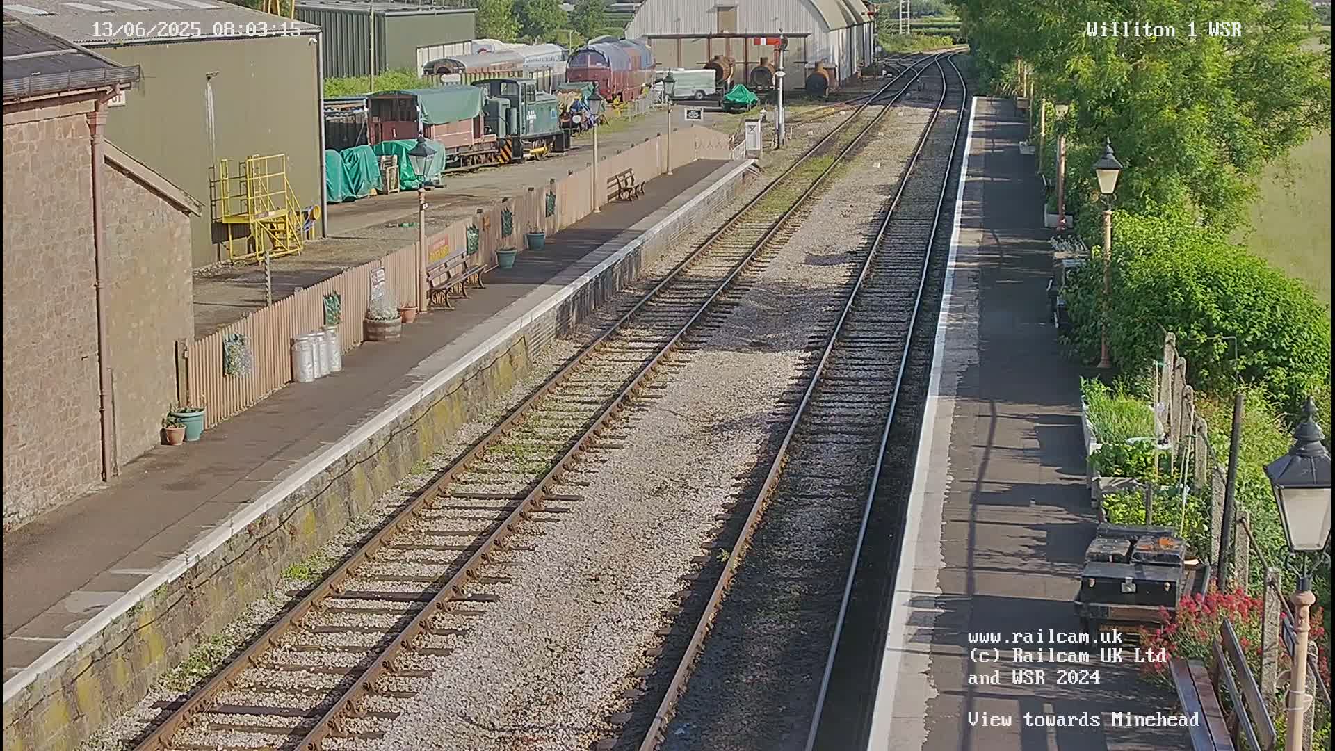 A sunny day at a train station shows a view down two parallel train tracks, with railway cars and buildings visible in the background.