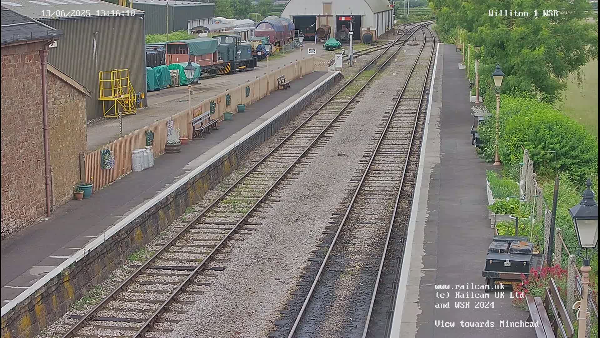 A train station platform with multiple train tracks, railway cars, and a building in the background on a partly sunny day.