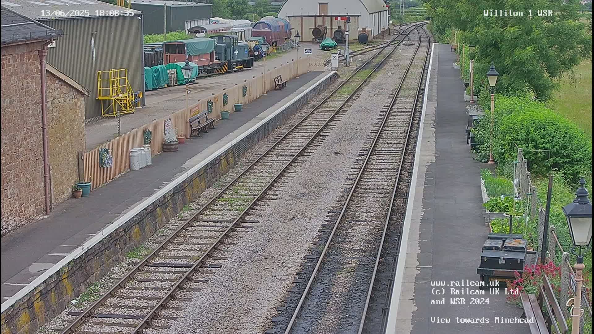 A railway station platform with multiple train tracks, several train cars visible in the background, and a garden alongside the platform on a partly sunny day.
