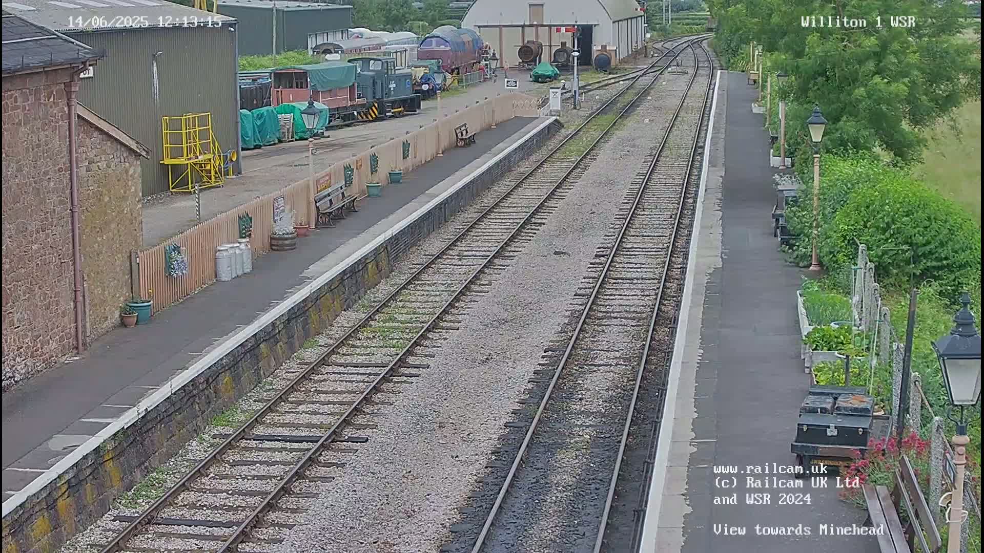 A train station platform with multiple railway tracks stretches into the distance on a partly sunny day,  with various railcars and buildings visible nearby.