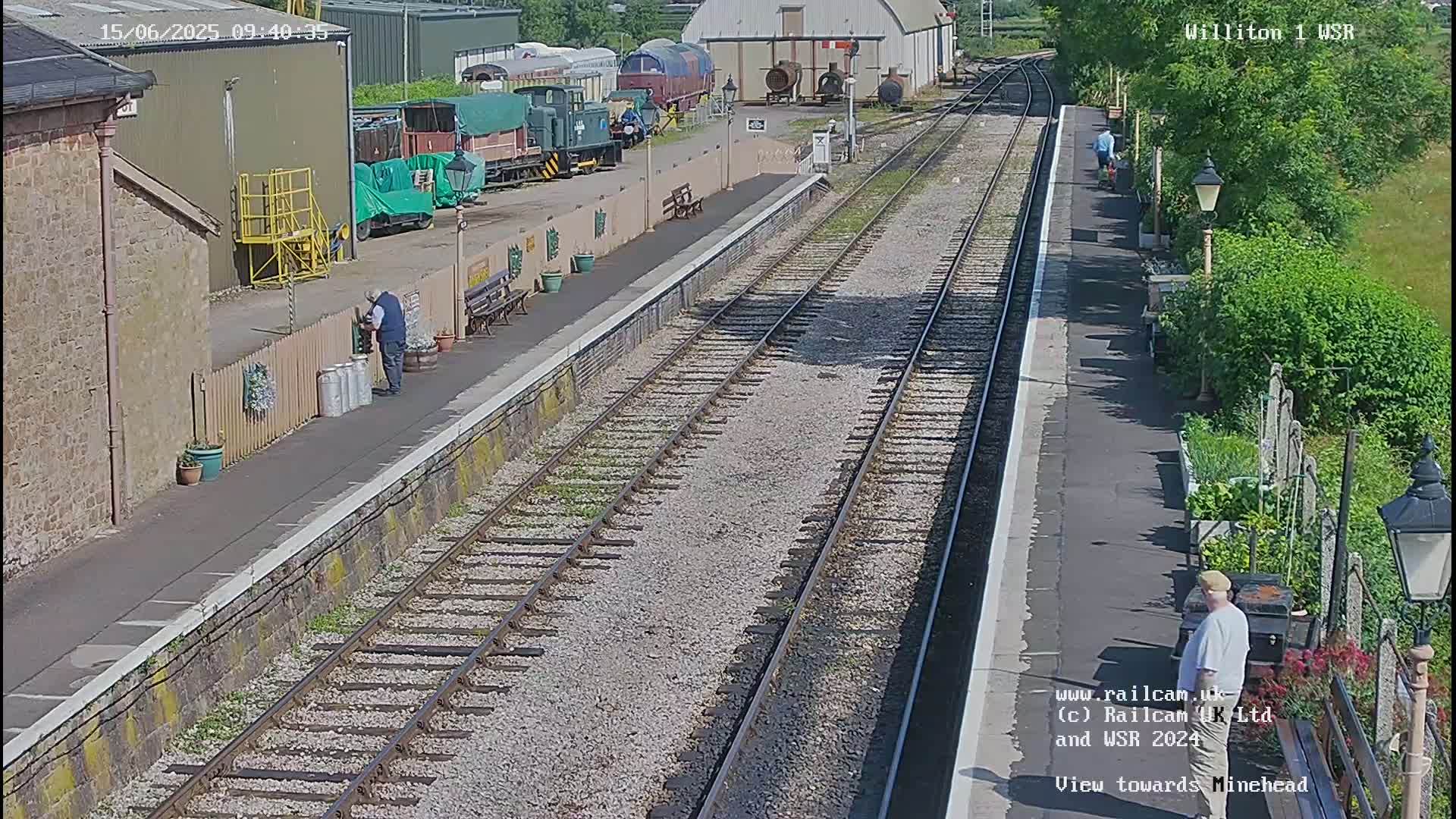A sunny day at a train station with multiple train tracks, some railway cars, and a few people.