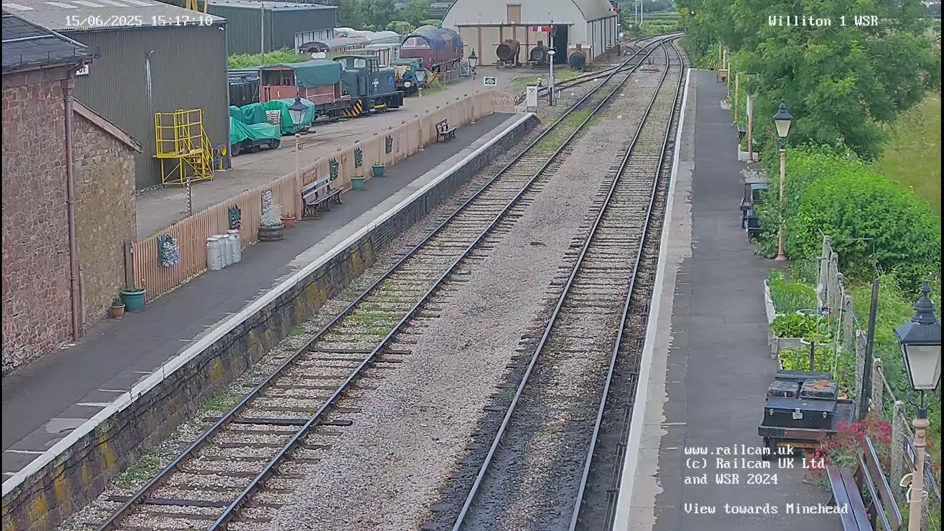 A railway station platform with multiple train tracks, railway cars, and a small garden is visible on a partly sunny day.