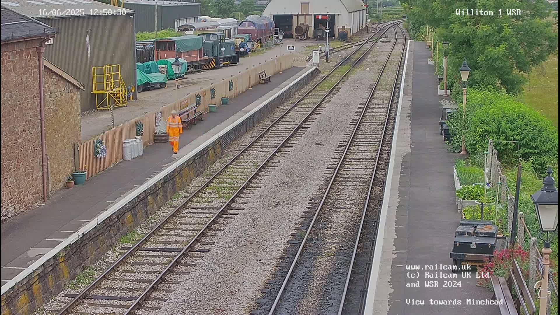 A person in an orange work jumpsuit walks along a railway platform next to train tracks, with several railway cars and buildings visible in the background, on an overcast day.