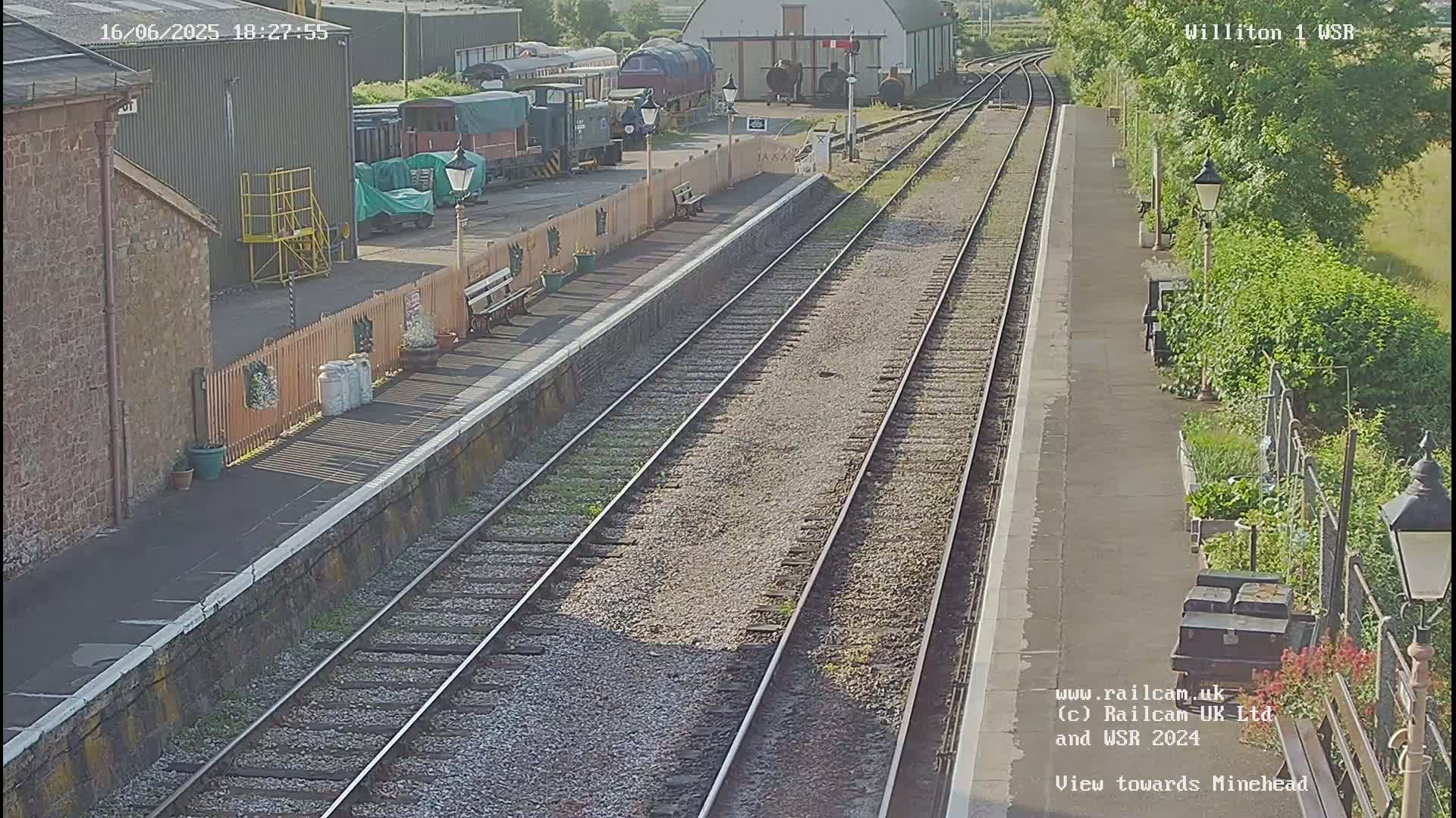 A sunny view of a train station platform with railway tracks extending into the distance, featuring several parked trains and railway buildings.