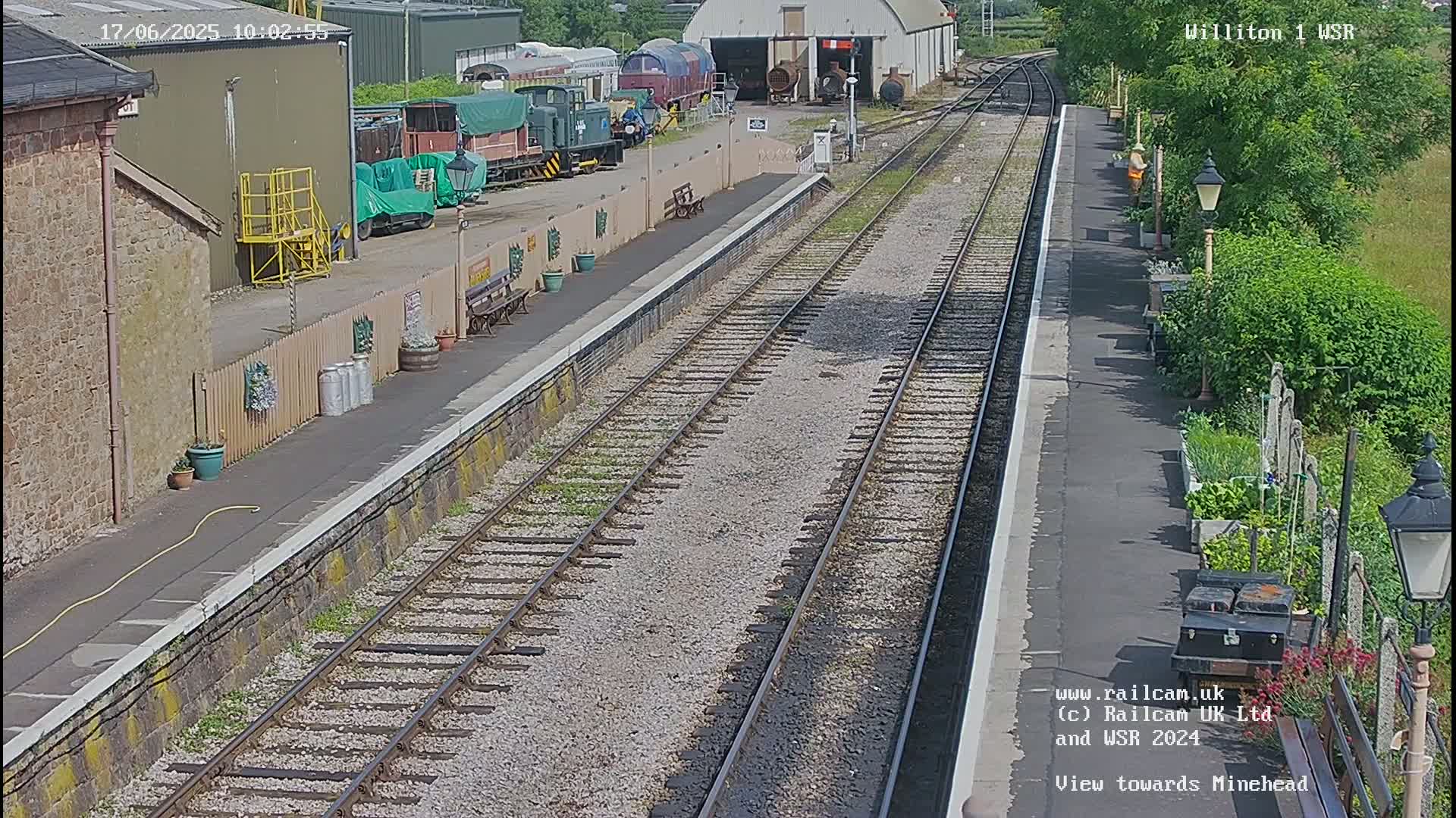 A sunny view of a railway station platform with multiple train tracks, railway cars, and a garden alongside.