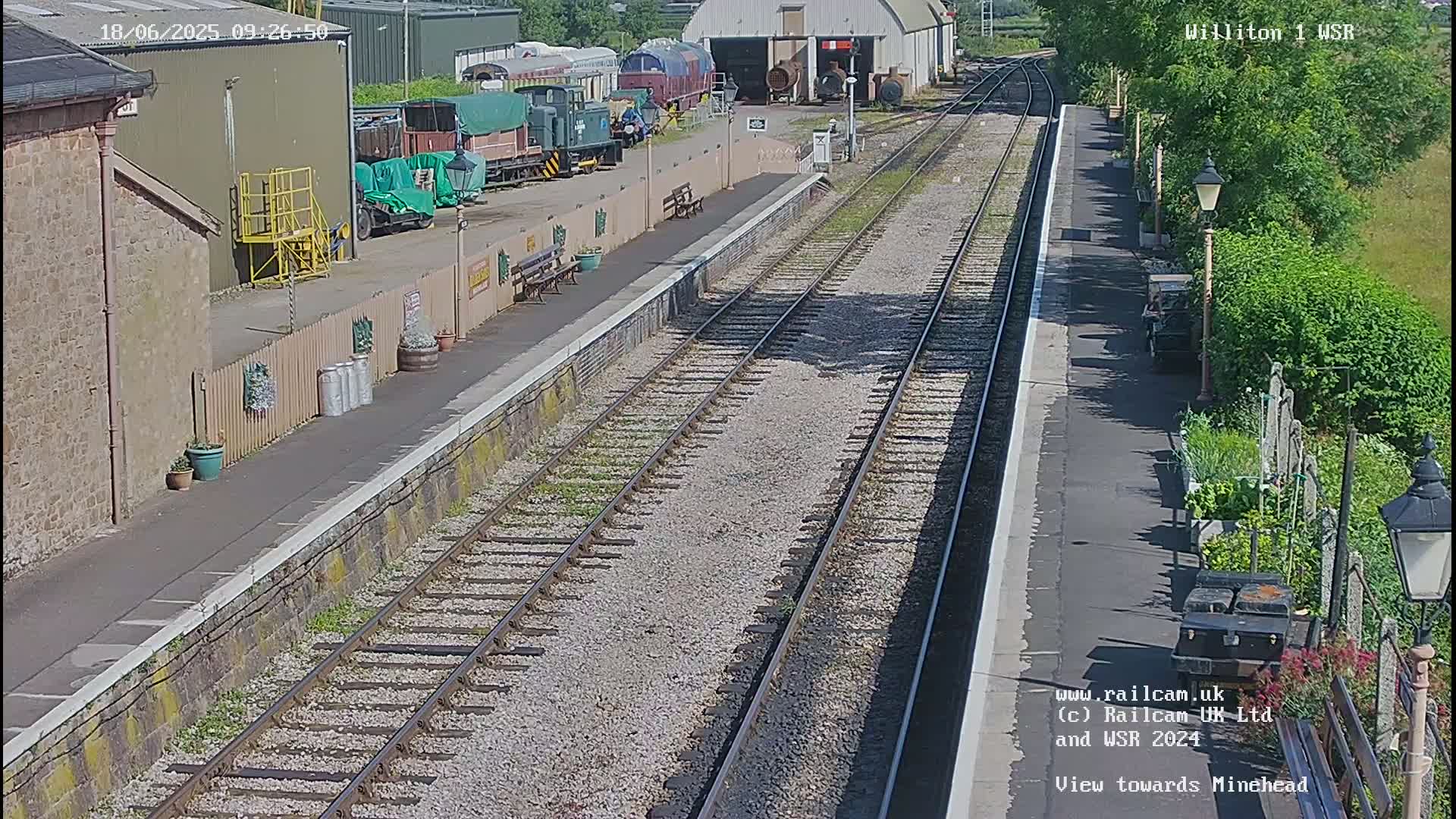 A sunny view of a train station platform with railway tracks extending into the distance, several train cars parked nearby, and a small garden beside the platform.