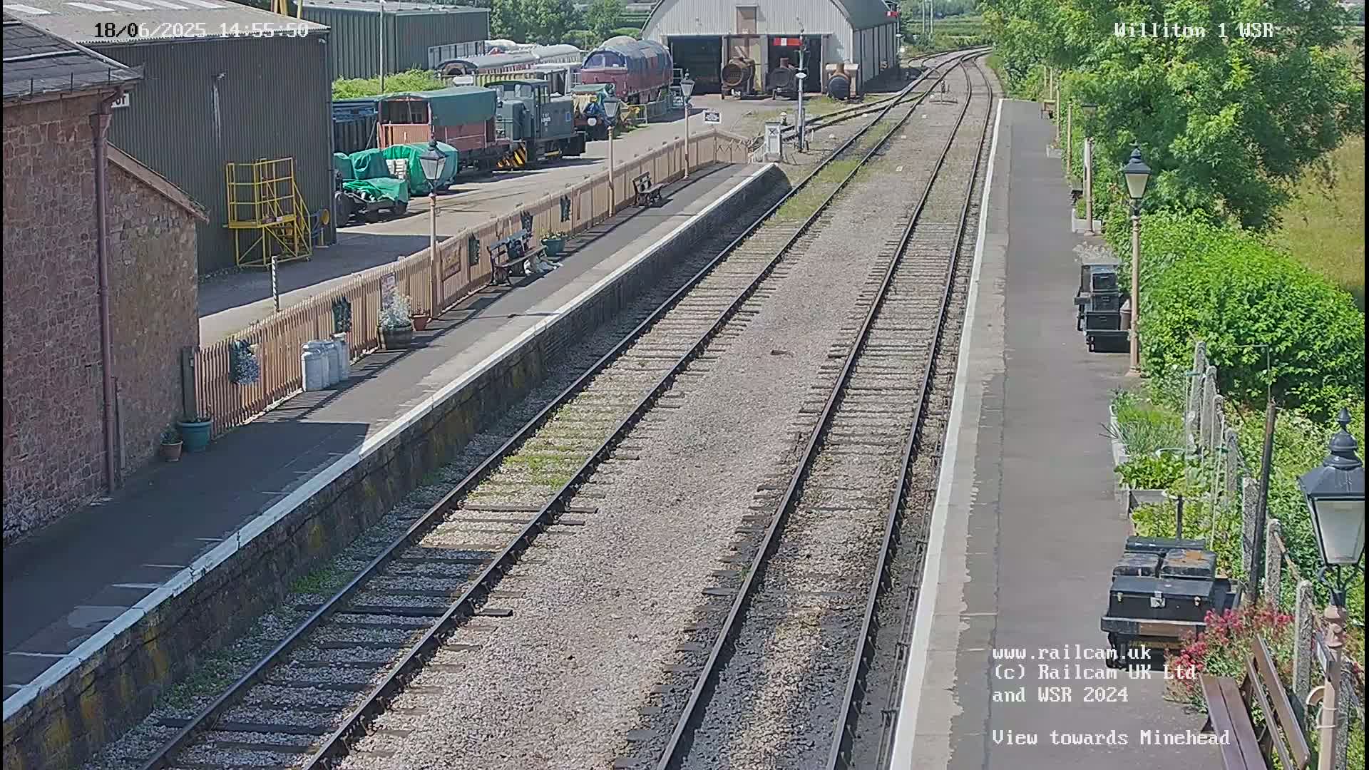 A sunny view of a train station platform with multiple train tracks extending into the distance, alongside a rail yard with several trains and train cars parked nearby.