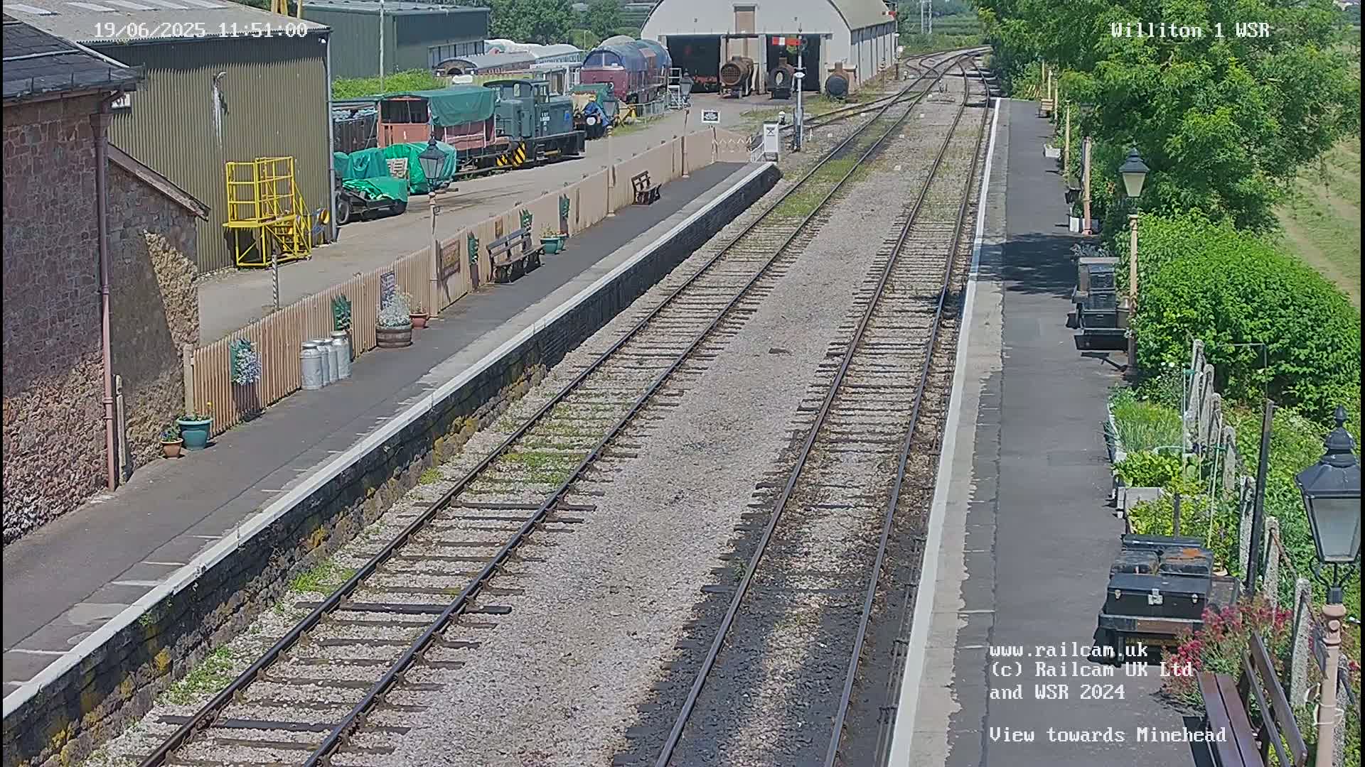 A sunny view of a train station platform with multiple railway tracks extending into the distance, showing several train cars and a depot in the background.